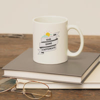 Ceramic coffee mug with 'Rise, Shine, Caffeinate' design, placed on books and glasses, ideal for coffee lovers.