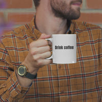 Man holding white coffee mug with 'Drink coffee' text, wearing plaid shirt in cozy setting.