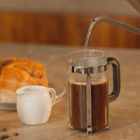 Hot water being poured into a French press with coffee, accompanied by bread and a milk jug on a kitchen countertop.