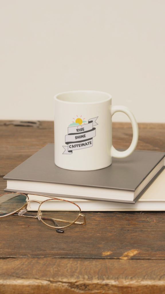 Ceramic coffee mug with 'Rise, Shine, Caffeinate' design, placed on books and glasses, ideal for coffee lovers.