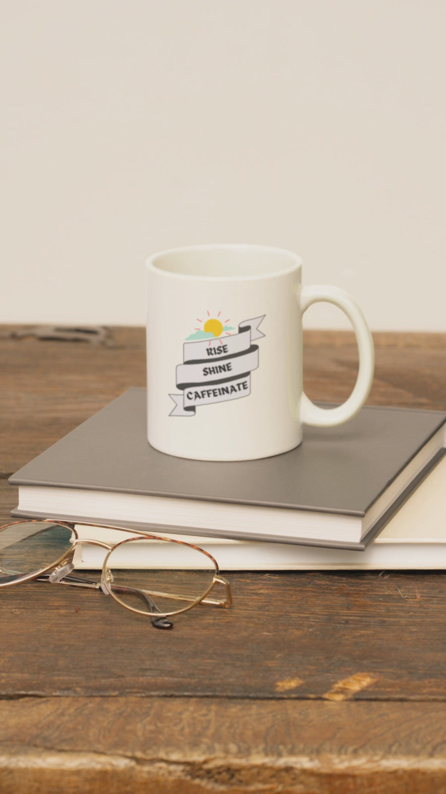 Ceramic coffee mug with 'Rise, Shine, Caffeinate' design, placed on books and glasses, ideal for coffee lovers.