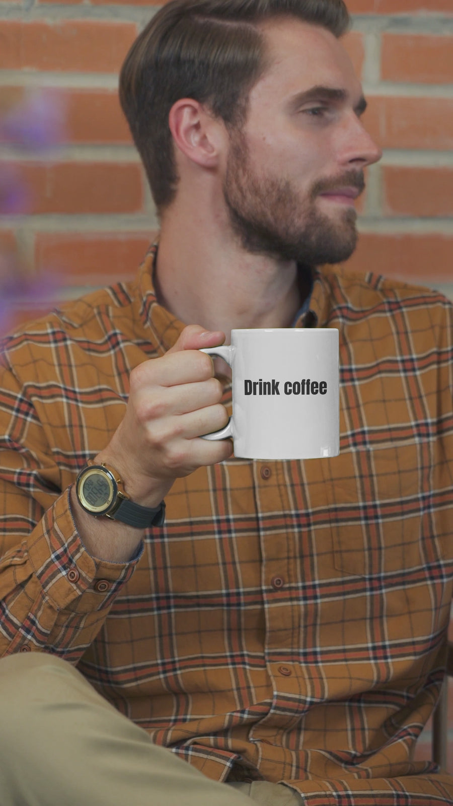Man holding white coffee mug with 'Drink coffee' text, wearing plaid shirt in cozy setting.
