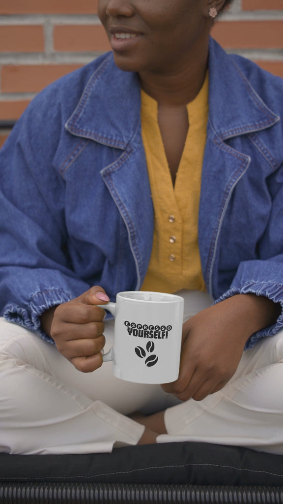 Person wearing a denim jacket holding a coffee mug with 'Espresso Yourself' text and coffee bean design.