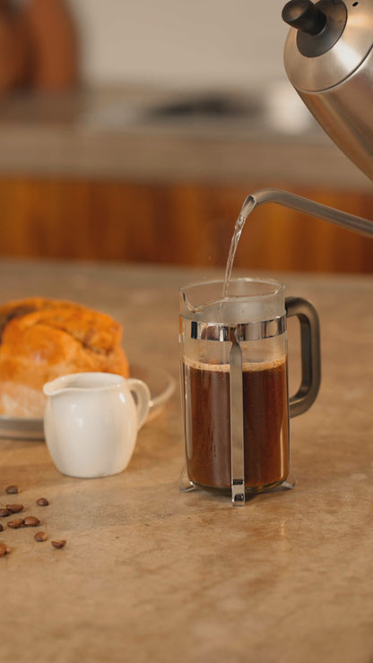 Hot water being poured into a French press with coffee, accompanied by bread and a milk jug on a kitchen countertop.