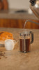 Hot water being poured into a French press with coffee, accompanied by bread and a milk jug on a kitchen countertop.